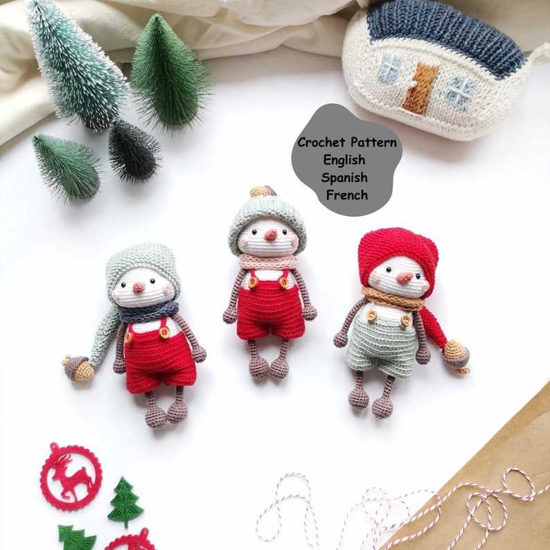 May include: Three handmade crochet dolls featuring white faces, pink cheeks, and small noses. Each doll wears a unique hat, scarf, and overalls with buttons. A gray speech bubble contains the text: Crochet Pattern English Spanish French.