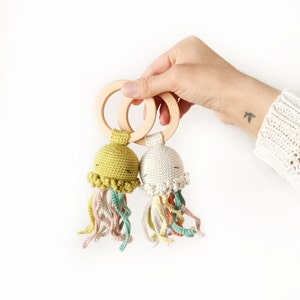 May include: Two crocheted jellyfish toys attached to smooth, round wooden rings. One jellyfish is olive green, and the other is off-white. Both feature embroidered closed eyes and multiple dangling, curly tentacles in muted shades of pink, yellow, and teal.