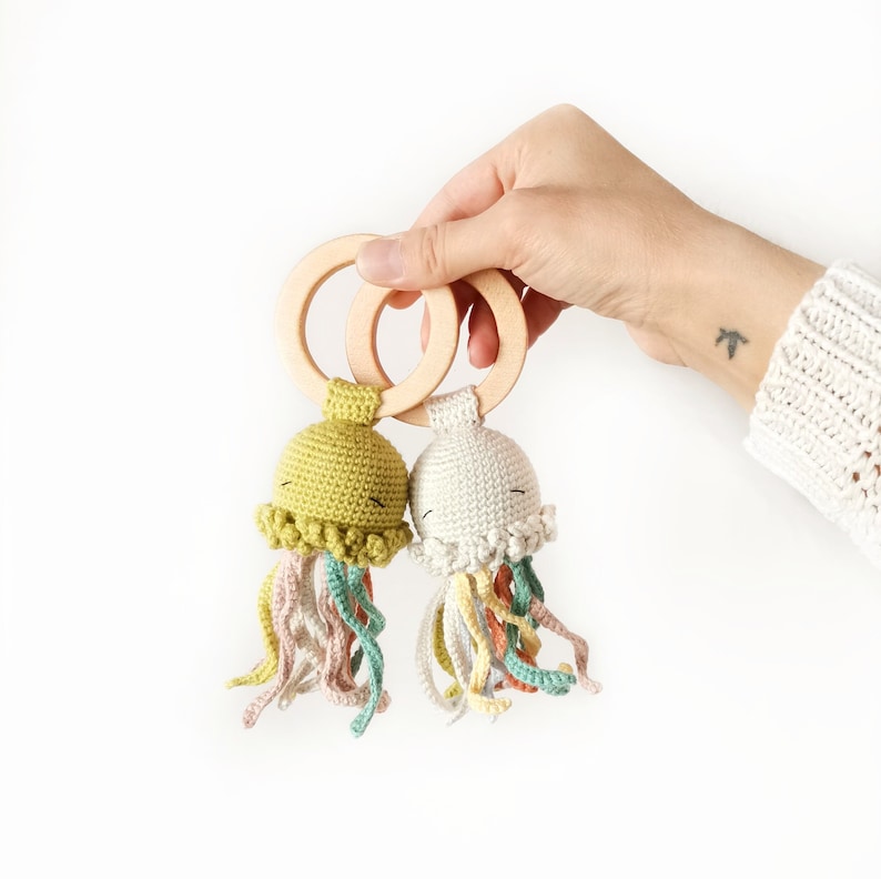 May include: Two crocheted jellyfish toys attached to smooth, round wooden rings. One jellyfish is olive green, and the other is off-white. Both feature embroidered closed eyes and multiple dangling, curly tentacles in muted shades of pink, yellow, and teal.