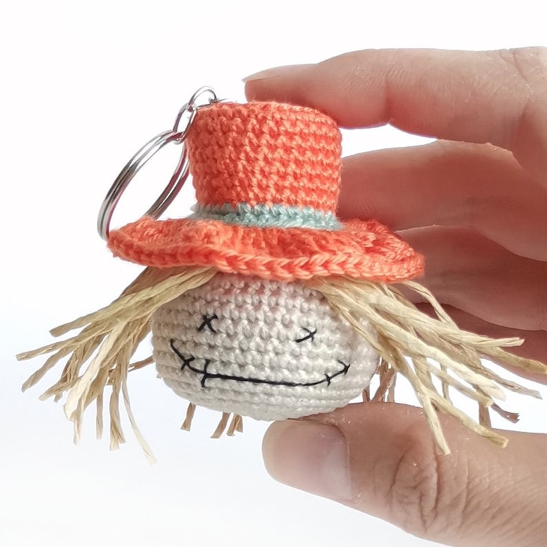 May include: A crochet keychain featuring a round, off-white head with stitched black X-shaped eyes and a stitched mouth. The figure wears a bright orange hat with a light blue band and has straw-like hair protruding from beneath the brim.