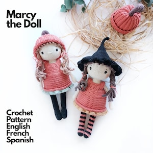 May include: Two handmade crochet dolls labelled Marcy the Doll. One doll wears a pink hat and dress with black legs. The other wears a black pointed hat and pink dress with striped legs. A separate pink crochet pumpkin is included in the display.