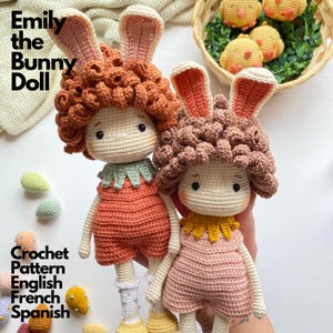 May include: Two handmade crochet dolls featuring bunny ears, curly brown hair, and outfits in orange and pink. The dolls have embroidered facial features and wear ruffled collars. The text Emily the Bunny Doll Crochet Pattern English French Spanish is visible on the left side of the image.