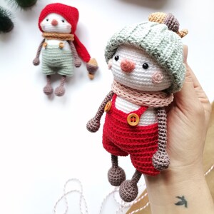 May include: Two handmade crochet dolls featuring white round heads, small black eyes, and orange noses. One doll wears a red hat, a tan scarf, and sage green overalls. The other doll wears a sage green ribbed hat, a pink scarf, and red overalls.