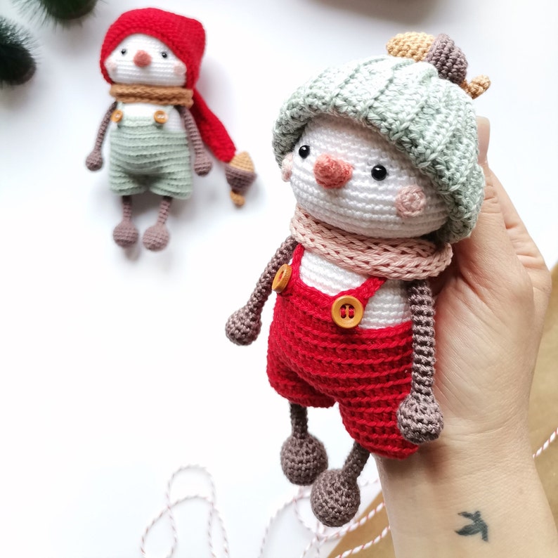 May include: Two handmade crochet dolls featuring white round heads, small black eyes, and orange noses. One doll wears a red hat, a tan scarf, and sage green overalls. The other doll wears a sage green ribbed hat, a pink scarf, and red overalls.