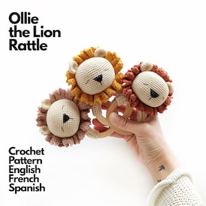 Op de afbeelding: Drie Ollie the Lion rammelaar haakspeeltjes met beige gezichten, zwarte geborduurde ogen en neuzen, omringd door manen in mosterdgeel, terracotta en oudroze. Elke leeuwenkop is bevestigd aan een gladde, ronde houten ring als handvat.