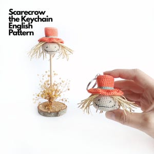 Peut inclure: Un porte-clés épouvantail en crochet avec un chapeau pêche, des cheveux en paille et des traits de visage brodés. Le texte 'Scarecrow the Keychain English Pattern' est visible. Un épouvantail est sur un support en bois, l'autre est tenu.