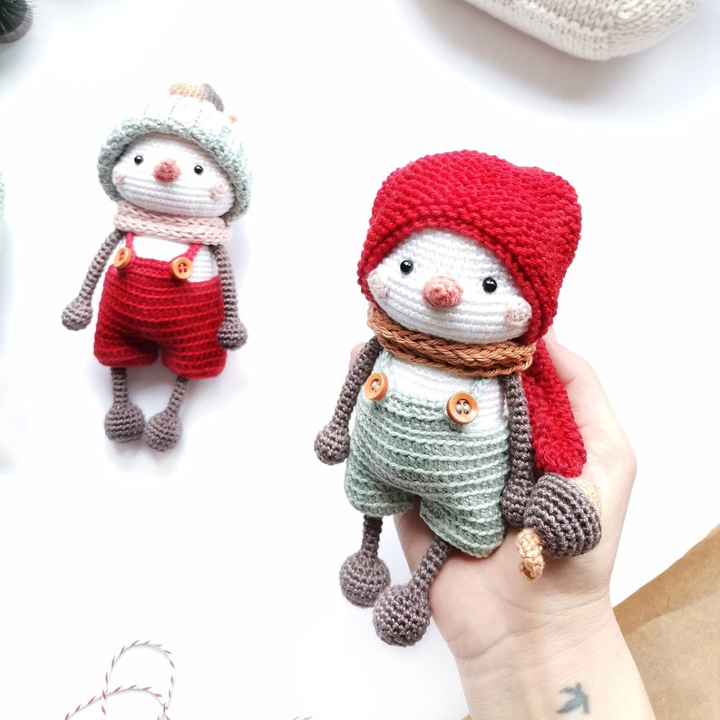 May include: Two handmade crochet dolls featuring round white heads with small black eyes and pink cheeks. One doll wears a red knit hat, brown scarf, and sage green overalls with wooden buttons. The other wears a mint hat, pink scarf, and red overalls.