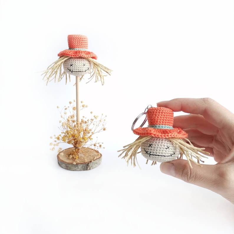 May include: A crochet scarecrow head with a beige face, stitched X-shaped eyes, a stitched mouth, and straw-like hair. It wears an orange hat with a light blue band. One version is on a wooden stick, and the other is a keychain with a metal ring.