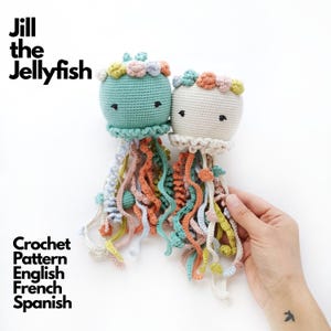 Puede incluir: Dos medusas de ganchillo, una de color verde menta y otra crema, cada una con una corona de pequeñas flores pastel, ojos bordados y largos tentáculos rizados de varios colores. El texto dice: Jill the Jellyfish Crochet Pattern English French Spanish.