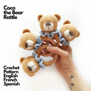 May include: Three crocheted bear rattles with tan yarn heads, white muzzles, and dark brown embroidered noses. Each bear features a light blue ruffled collar attached to a smooth, round wooden ring. Text reads: Coco the Bear Rattle, Crochet Pattern English French Spanish.