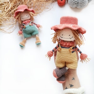 May include: Two handmade crochet scarecrow dolls featuring yarn hair, stitched facial features, and wide-brimmed hats. One doll wears teal overalls, while the other wears yellow overalls. Both dolls have striped long-sleeve shirts and brown boots.