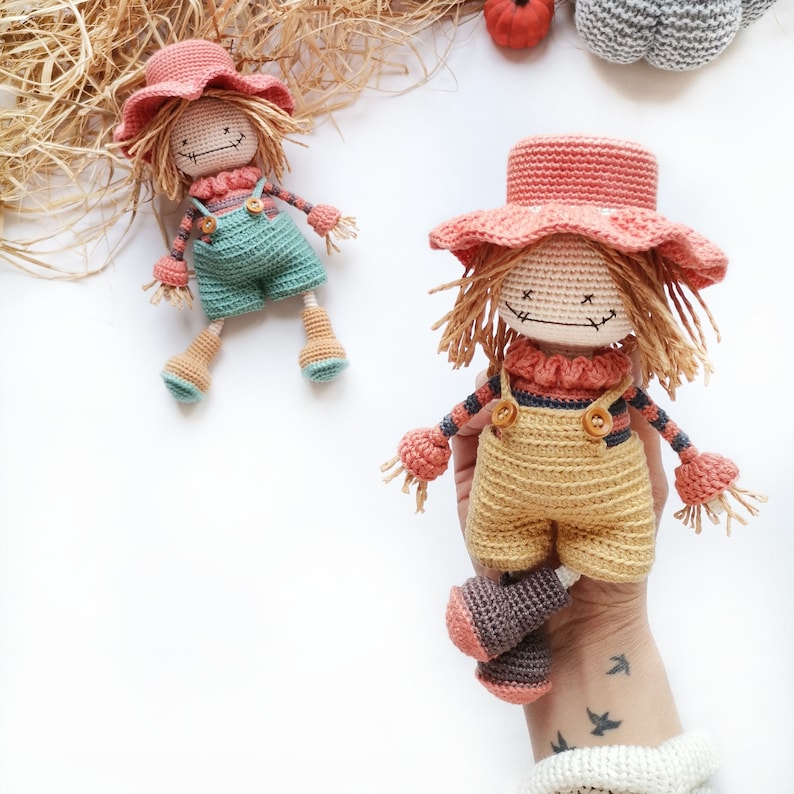 May include: Two handmade crochet scarecrow dolls featuring yarn hair, stitched facial features, and wide-brimmed hats. One doll wears teal overalls, while the other wears yellow overalls. Both dolls have striped long-sleeve shirts and brown boots.