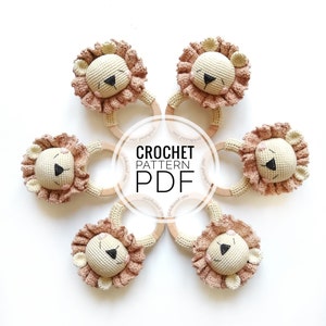 May include: Five crochet lion rattles arranged in a circle, each featuring a cream-colored head, a textured dusty pink mane, and a smooth wooden ring. The center of the image displays the text CROCHET PATTERN PDF in bold black lettering.