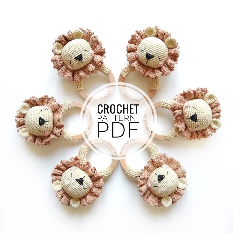 May include: Five crochet lion rattles arranged in a circle, each featuring a cream-colored head, a textured dusty pink mane, and a smooth wooden ring. The center of the image displays the text CROCHET PATTERN PDF in bold black lettering.