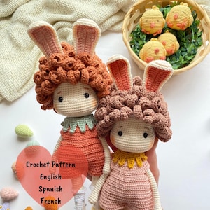 May include: Two handmade crochet dolls with bunny ears and curly hair, wearing dresses. One doll is wearing an orange dress with a green collar, and the other is wearing a pink dress with a yellow collar. The dolls are next to a basket of yellow crochet chicks. Text on the image says "Crochet Pattern English Spanish French".