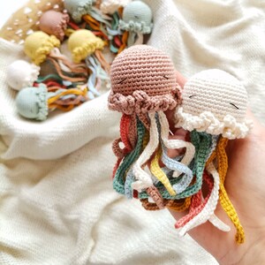 May include: Two small, handmade crochet jellyfish toys with rounded heads and dangling, multi-colored tentacles in shades of dusty pink, cream, mustard yellow, light blue, and coral. Each jellyfish features embroidered closed eyes.