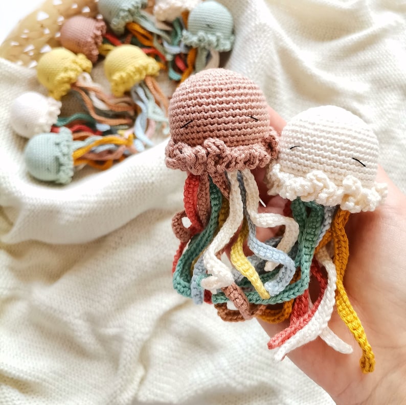 May include: Two small, handmade crochet jellyfish toys with rounded heads and dangling, multi-colored tentacles in shades of dusty pink, cream, mustard yellow, light blue, and coral. Each jellyfish features embroidered closed eyes.