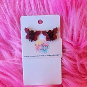 May include: A pair of dark red moth-shaped earrings on a white card. The earrings have a glittery texture and are set against a vibrant pink, fluffy background. The card features the text "YaliPop Designs" and a website address.