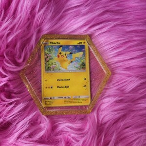 May include: A yellow Pikachu Pokémon trading card inside a gold glitter resin hexagon. The card features a yellow Pikachu character with the text "Quick Attack" and "Electro Ball". The background is a vibrant pink, fluffy surface.