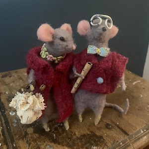May include: Two gray felt mice wearing clothing. The mouse on the left is wearing a red coat, a floral necklace, and a white lace bag. The mouse on the right is wearing a blue and white patterned jacket, a bow tie, and round silver glasses.