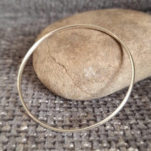 May include: A plain, silver bangle bracelet. The thin, circular bracelet is shown on a textured, grey surface with a large, smooth, brown rock in the background. The bracelet has a minimalist design.