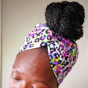 May include: A woman with dark hair styled in a bun is wearing a white headband with a colorful leopard print. The headband is tied in a knot at the top of her head. She is wearing large hoop earrings and a black shirt.