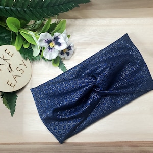 May include: A navy blue headband with a gold geometric pattern. The headband is twisted at the center. The image also includes a wooden disc with the words "TAKE IT EASY" and some greenery.