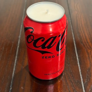 May include: A red Coca-Cola can candle with a white wax top and a black wick. The can has the text "Coca-Cola Zero Sugar" and "Zero Calorie" printed on it.