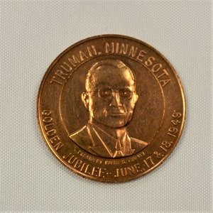 May include: A copper-colored coin with the text "TRUMAN MINNESOTA GOLDEN JUBILEE JUNE 17 & 18, 1949" around the edge. The center of the coin features a portrait of a man wearing glasses and a suit.