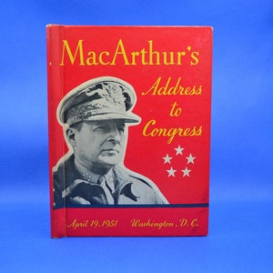 Op de afbeelding: Een rood boek met de titel "MacArthur's Address to Congress" in gouden letters. Op de omslag staat een zwart-witfoto van generaal Douglas MacArthur in zijn militaire uniform. Het boek is uitgegeven op 19 april 1951 in Washington, D.C.