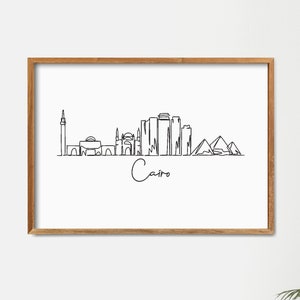 Może przedstawiać: Czarno-biały rysunek liniowy panoramy Kairu, w tym piramidy, meczet i wysokie budynki. Rysunek jest oprawiony w jasną drewnianą ramę i wisi na białej ścianie.