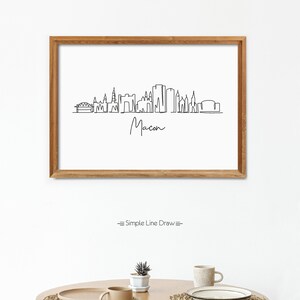 Puede incluir: Un dibujo en blanco y negro de la l&iacute;nea del horizonte de Macon, Georgia. El dibujo est&aacute; enmarcado en un marco de madera marr&oacute;n claro y est&aacute; colgado en una pared blanca. El texto "Simple Line Draw" est&aacute; debajo del marco.