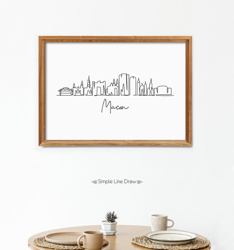Puede incluir: Un dibujo en blanco y negro de la l&iacute;nea del horizonte de Macon, Georgia. El dibujo est&aacute; enmarcado en un marco de madera marr&oacute;n claro y est&aacute; colgado en una pared blanca. El texto "Simple Line Draw" est&aacute; debajo del marco.