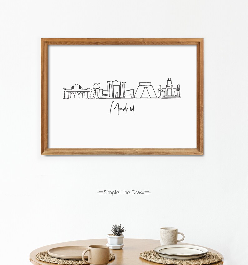 May include: A framed print featuring a minimalist line drawing of the Madrid skyline in black against a white background. The word "Madrid" is written below the skyline in cursive. The frame is made of light brown wood.