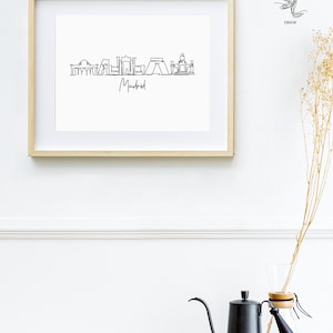 May include: A framed print featuring a minimalist line drawing of the Madrid skyline in black against a white background. The word "Madrid" is written below the skyline in a handwritten style. The frame is light-colored wood.