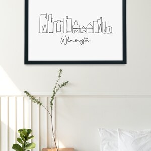 May include: Black and white line art print of the Wilmington skyline with the text "Wilmington" below the skyline. The print is framed in a black frame and hanging on a white wall.