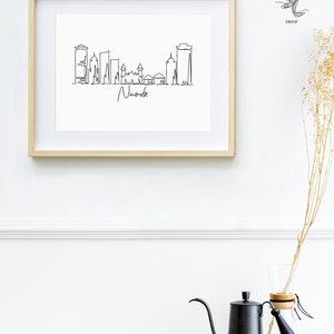 Puede incluir: Un dibujo lineal en blanco y negro enmarcado del horizonte de Nairobi. La obra de arte est&aacute; en un marco de madera clara y presenta el nombre de la ciudad en escritura cursiva. La imagen se muestra en una pared blanca sobre una superficie de madera.