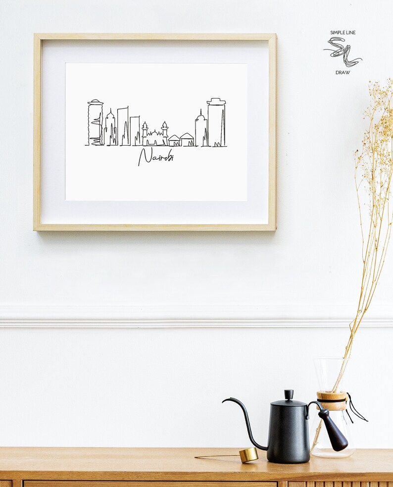 Puede incluir: Un dibujo lineal en blanco y negro enmarcado del horizonte de Nairobi. La obra de arte est&aacute; en un marco de madera clara y presenta el nombre de la ciudad en escritura cursiva. La imagen se muestra en una pared blanca sobre una superficie de madera.