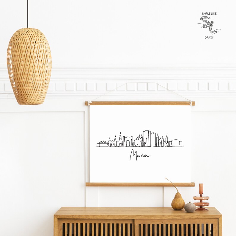 Puede incluir: Un dibujo en blanco y negro de la l&iacute;nea del horizonte de Macon, Georgia. El dibujo est&aacute; en un lienzo blanco con un marco de madera. El lienzo est&aacute; colgado en una pared blanca sobre un gabinete de madera.