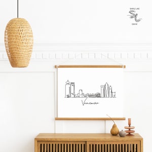 Puede incluir: Un dibujo en blanco y negro de la l&iacute;nea del horizonte de Vancouver con la palabra "Vancouver" escrita debajo. El dibujo est&aacute; enmarcado y colgado en una pared blanca sobre un armario de madera.