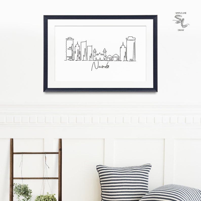 Puede incluir: Impresi&oacute;n art&iacute;stica enmarcada en blanco y negro del horizonte de Nairobi. La obra de arte presenta un dise&ntilde;o minimalista de edificios y el nombre de la ciudad en cursiva. La impresi&oacute;n se muestra en una pared blanca sobre una escalera decorativa y almohadas.