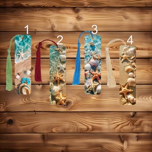 May include: Four rectangular bookmarks with beach-themed designs. Each bookmark features a different design of seashells, starfish, and sand, with a tassel at the top. The tassels are green, burgundy, blue, and beige.