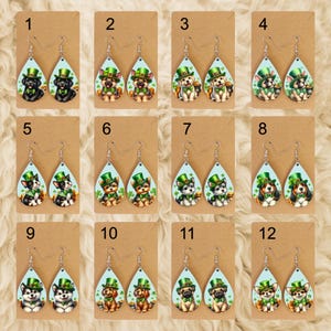 May include: A collection of twelve pairs of teardrop-shaped earrings. Each earring features a different dog breed wearing a green leprechaun hat and shamrock accents. The earrings are displayed on brown cardstock.