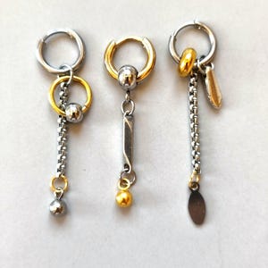 May include: Three hoop earrings featuring mixed metal designs. The first has a silver hoop, gold ring, silver chain, and silver bead. The second has a gold hoop, silver bar, and gold bead. The third has a silver hoop, gold bead, silver chain, and oval charm.