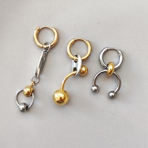 Op de afbeelding: Drie oorbellen van gemengde metalen met goud- en zilverkleurige onderdelen. De eerste is een ring met een hangende staaf en een kleine ring met twee kralen. De tweede is een ring met een gebogen staaf en een grote gouden kraal. De derde is een ring met een hoefijzervormige bedel.