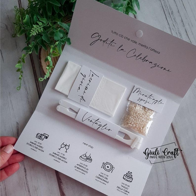 Op de afbeelding: Een witte kaart met de tekst "Goditi la Celebrazione" in zwarte schrijfletters. De kaart is dubbelgevouwen en bevat een wit papieren zakdoekje, een witte waaier, een klein zakje rijst en een witte kaart met de tekst "Lacrime di gioia" in zwarte schrijfletters. De kaart bevat ook een witte kaart met de tekst "Ventaglio" in zwarte schrijfletters. De kaart ligt op een wit oppervlak.