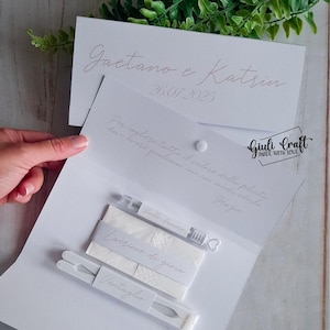 Op de afbeelding: Witte bruiloft gunst set met een witte kaart met de tekst "Gaetano e Katrin 26.07.2025" en "Per cogliere tutto il valore della felicita devi avere qualcuno con cui condividerla Grazie". De set bevat een klein flesje met bubbels, een pak tissues met het label "Lacrime di gioia" en een witte waaier met het label "Ventaglio".
