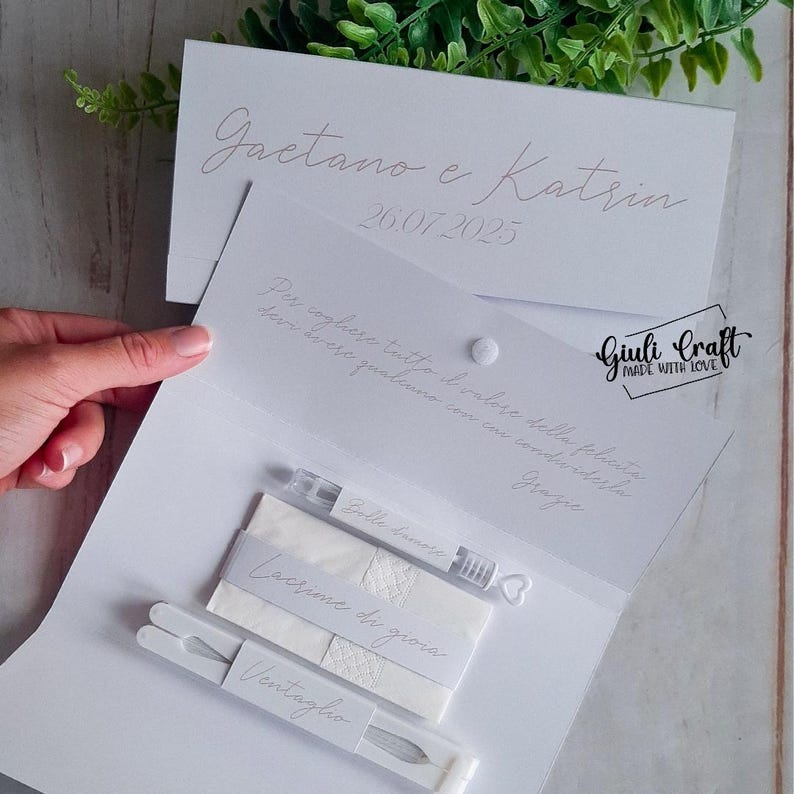Op de afbeelding: Witte bruiloft gunst set met een witte kaart met de tekst "Gaetano e Katrin 26.07.2025" en "Per cogliere tutto il valore della felicita devi avere qualcuno con cui condividerla Grazie". De set bevat een klein flesje met bubbels, een pak tissues met het label "Lacrime di gioia" en een witte waaier met het label "Ventaglio".