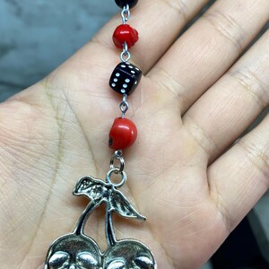 Puede incluir: Un dije colgante con un colgante de cerezas de metal plateado donde las cerezas tienen forma de calaveras. El dije est&aacute; unido a una cadena vertical de cuentas que incluye una bola ocho negra, una cuenta de calavera roja, un dado negro y otra calavera roja.
