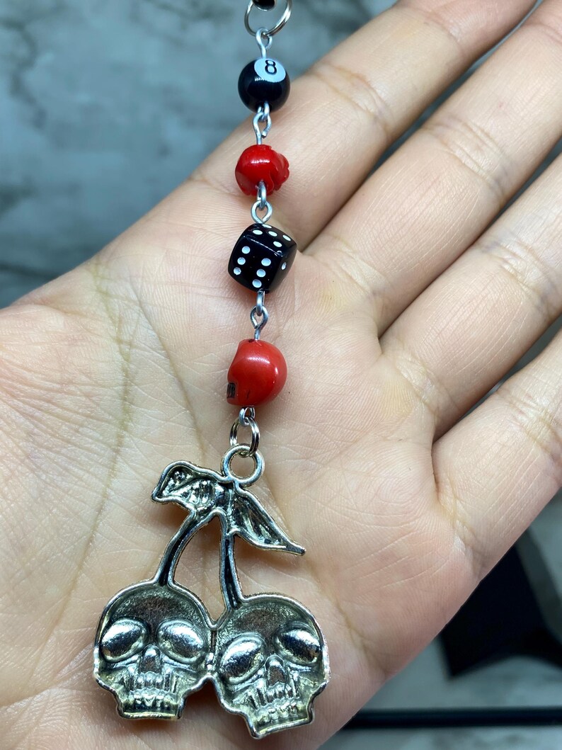 Puede incluir: Un dije colgante con un colgante de cerezas de metal plateado donde las cerezas tienen forma de calaveras. El dije est&aacute; unido a una cadena vertical de cuentas que incluye una bola ocho negra, una cuenta de calavera roja, un dado negro y otra calavera roja.